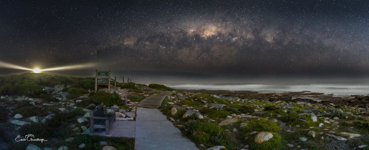 Milky Way above Kommetjie's rocky coastline, with a foreground path and a bright beam of light on the left horizon.