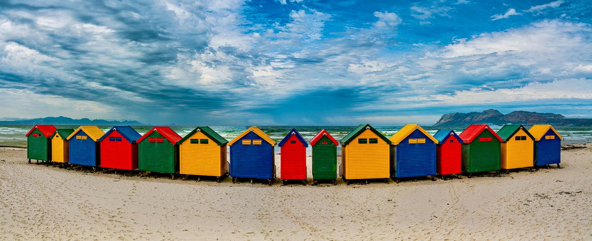 Wide panoramic view of colourful Muizenberg beach huts curving along the shoreline with False Bay, Muizenberg Peak and dramatic cloudscape behind