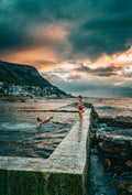 Father and son at Kalk Bay tidal pool at sunset with dramatic storm clouds and mountain backdrop, False Bay