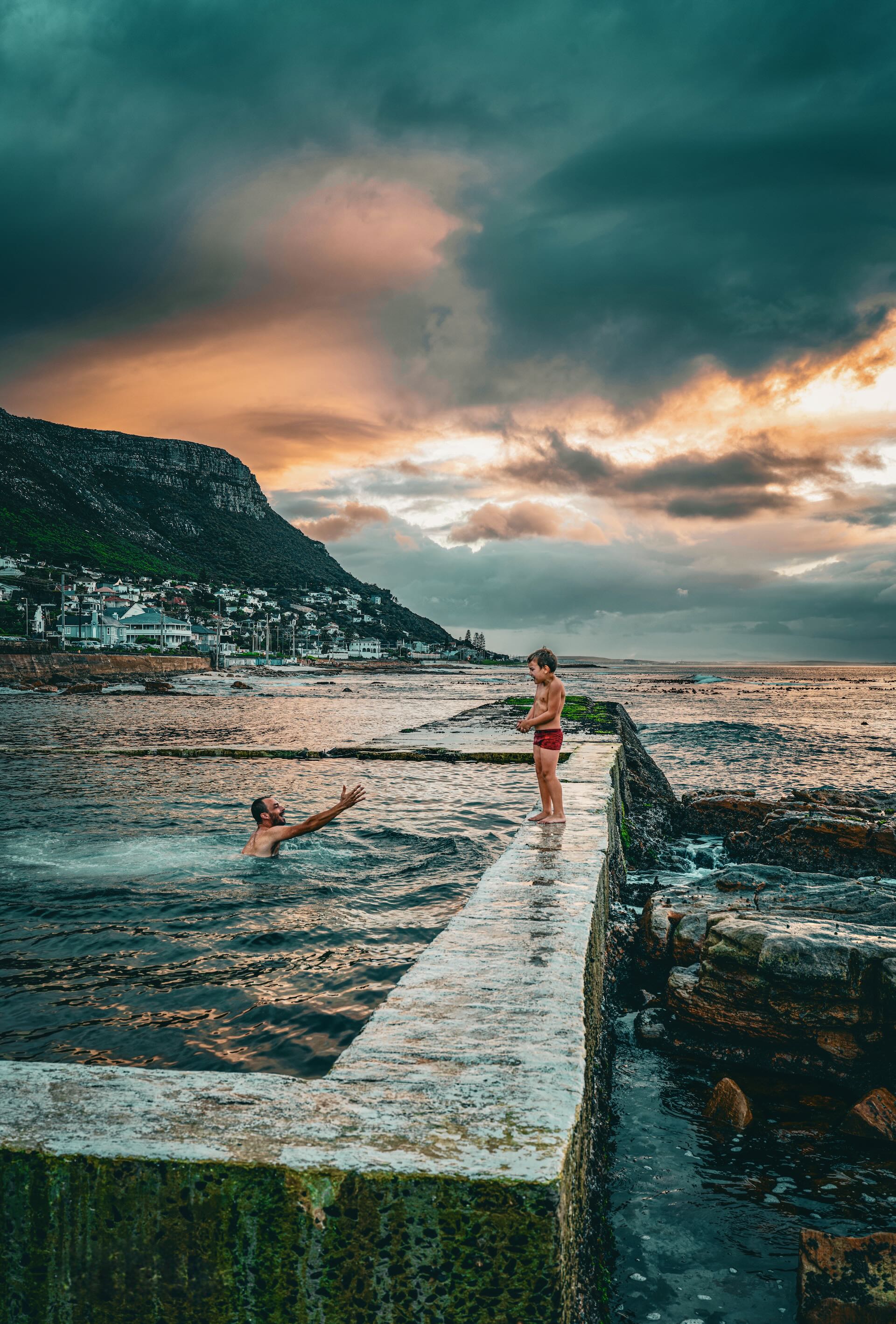 Father and son at Kalk Bay tidal pool at sunset with dramatic storm clouds and mountain backdrop, False Bay