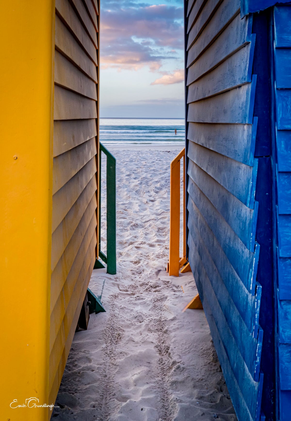 View through a narrow gap between yellow and blue Muizenberg beach huts looking toward the ocean at dusk with a lone figure on the beach