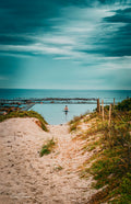 Lone swimmer in Glen Cairn tidal pool viewed from sandy dune path with turquoise False Bay beyond