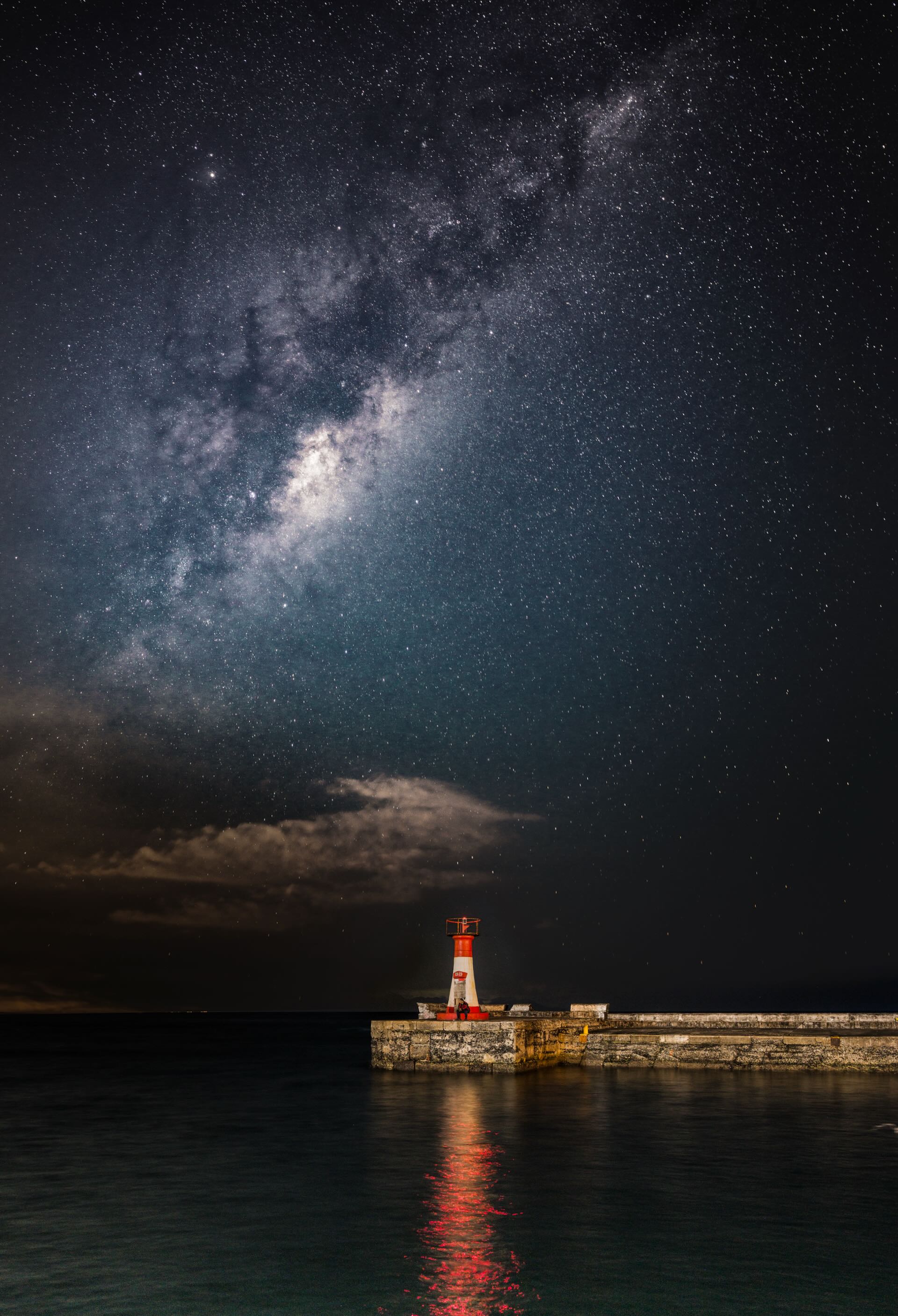 Kalk Bay harbour lighthouse glowing red under the Milky Way with reflection in still harbour water