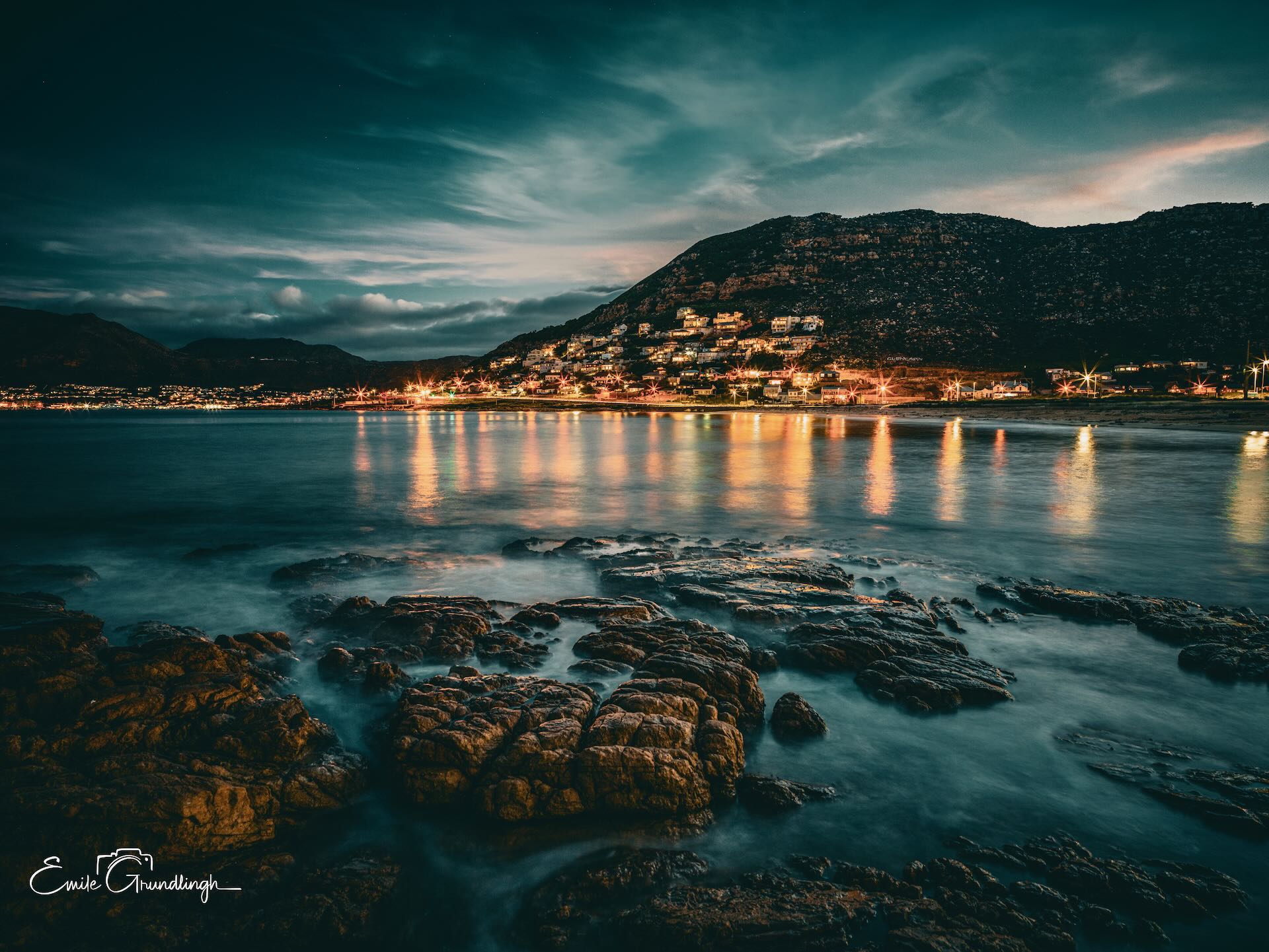 Glen Cairn coastal town lights reflecting on False Bay waters at night with rocky foreshore