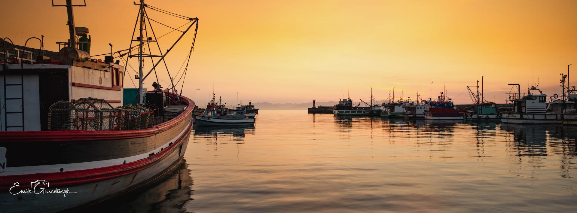 Kalk Bay fishing harbour at golden sunrise with boats moored in still glassy water and morning mist