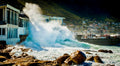 Massive wave crashing against Harbour House restaurant at Kalk Bay with fishing boats and mountain in background
