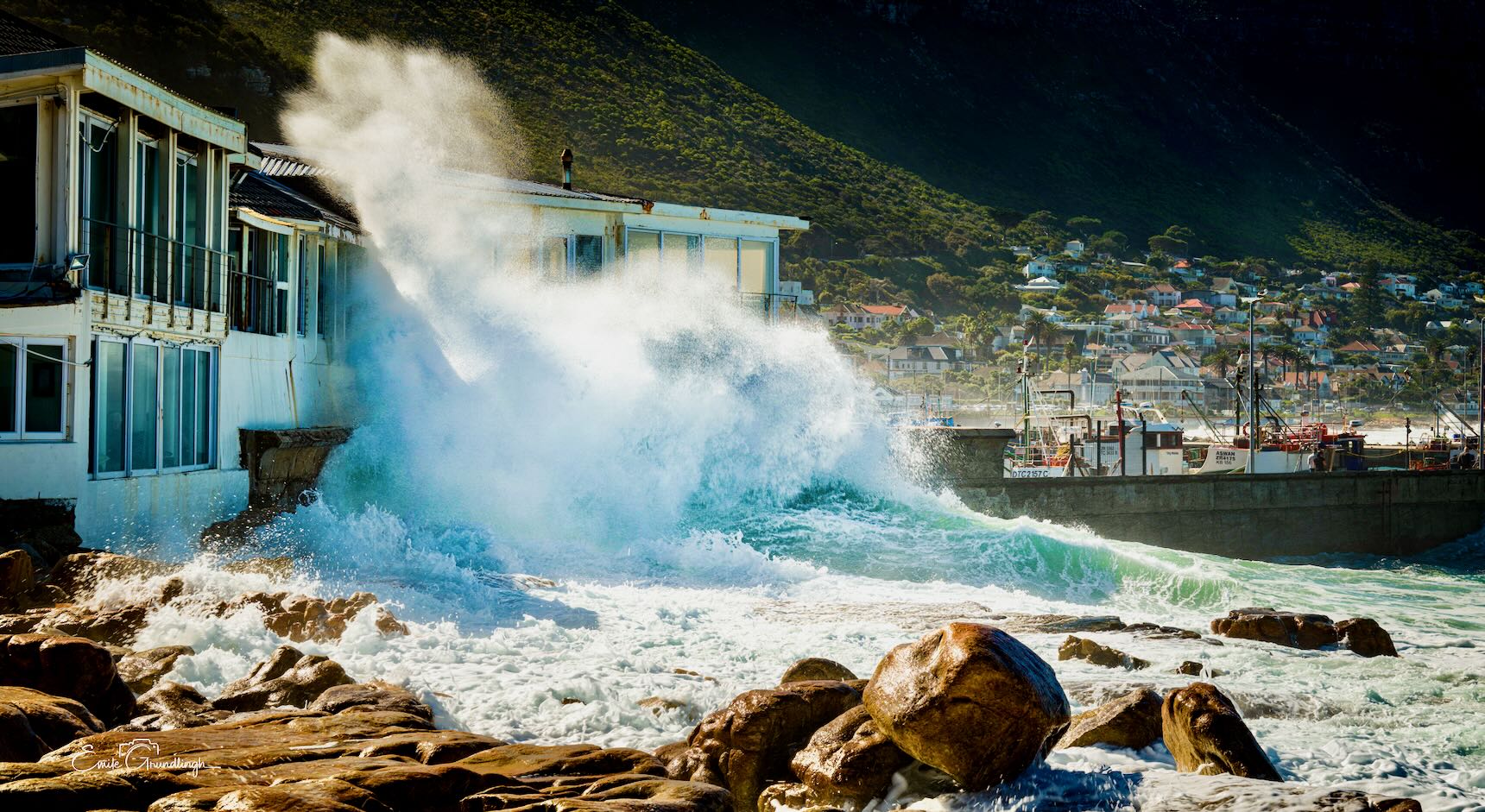 Massive wave exploding against old beachfront building at Kalk Bay Harbour with village and mountain behind