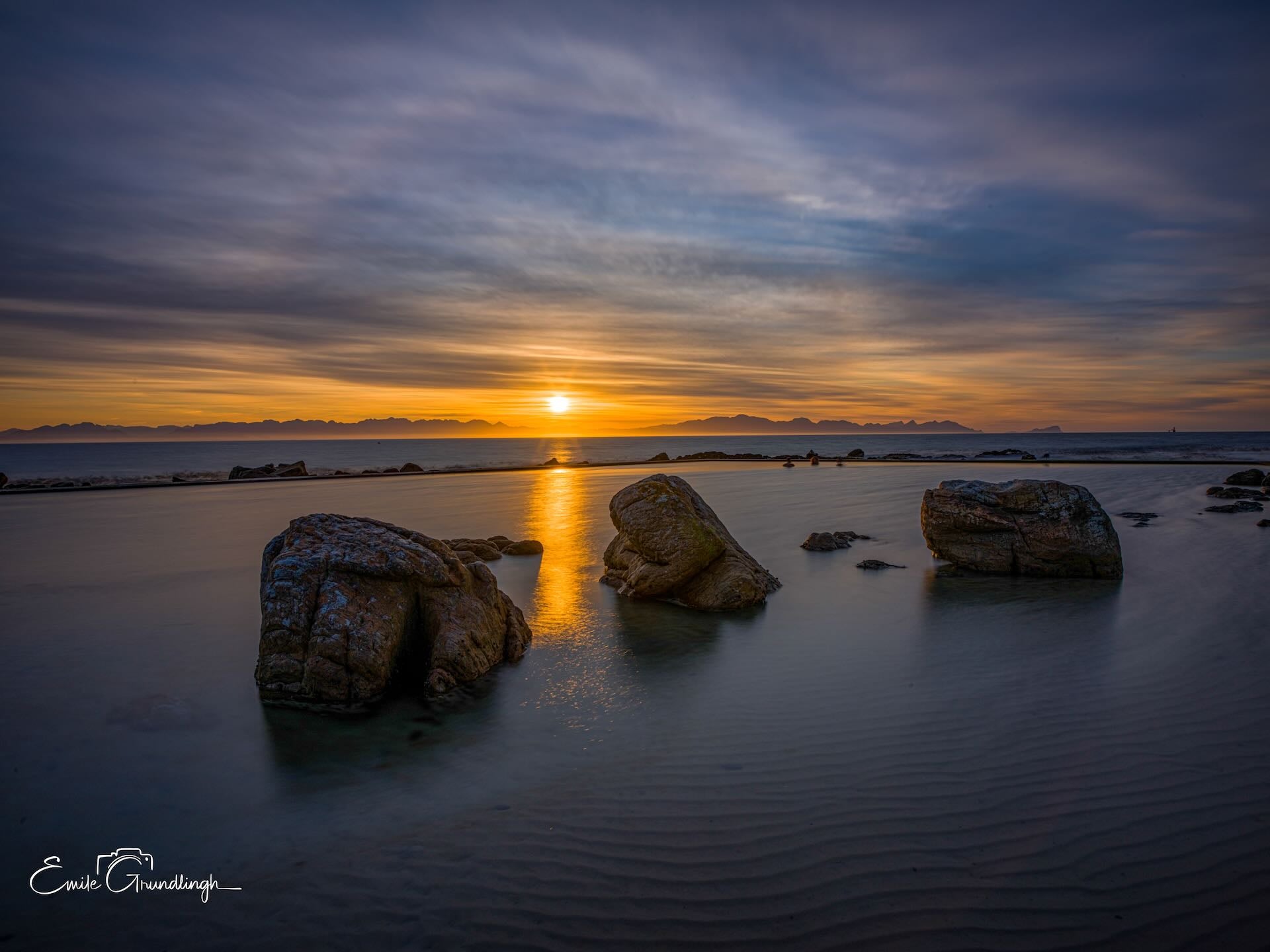 Three granite boulders in Glen Cairn tidal pool at sunset with golden reflection over False Bay