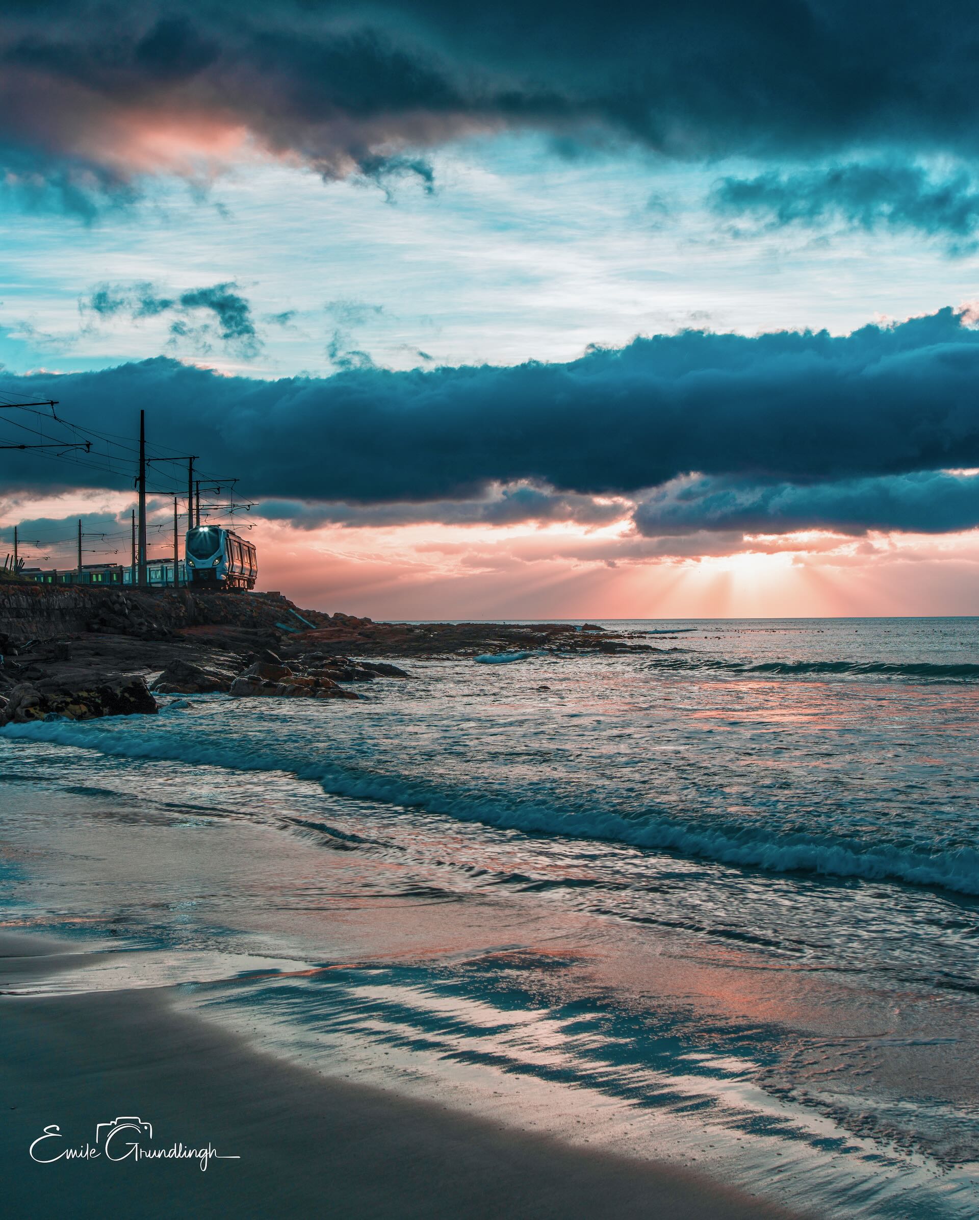 Metrorail train along the Glen Cairn coastline at sunset with dramatic storm clouds and golden sun rays over False Bay