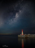 Red lighthouse on stone pier beneath a stunning Milky Way night sky reflecting on calm water