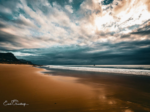 Wide golden Glen Cairn beach under dramatic teal and gold cloudscape with mountain headland, False Bay
