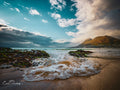 Low angle view from water's edge at Makriels Beach with mossy rocks, foamy waves and golden mountain under dramatic sky