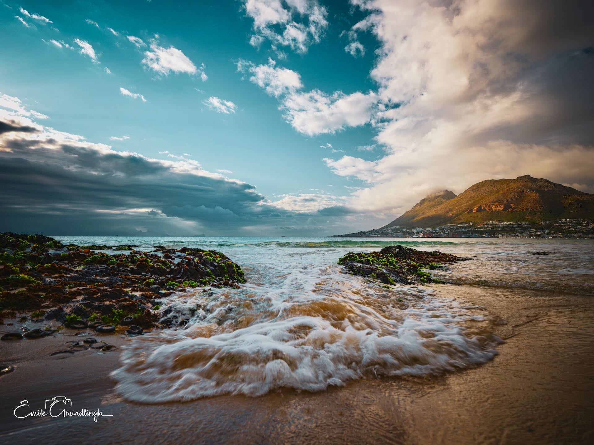 Low angle view from water's edge at Makriels Beach with mossy rocks, foamy waves and golden mountain under dramatic sky