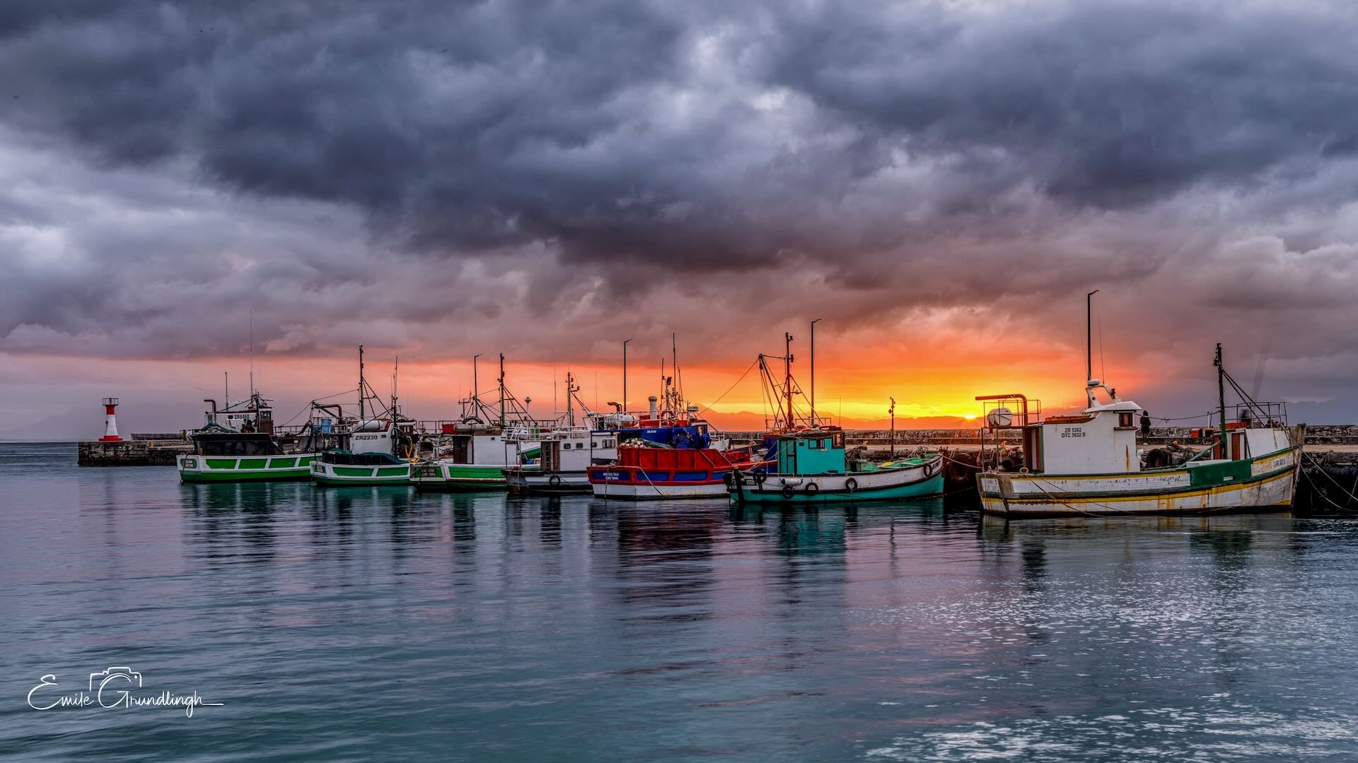 Kalk Bay fishing fleet silhouetted against blazing orange sunset with dramatic dark storm clouds overhead