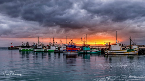 Kalk Bay fishing fleet silhouetted against blazing orange sunset with dramatic dark storm clouds overhead