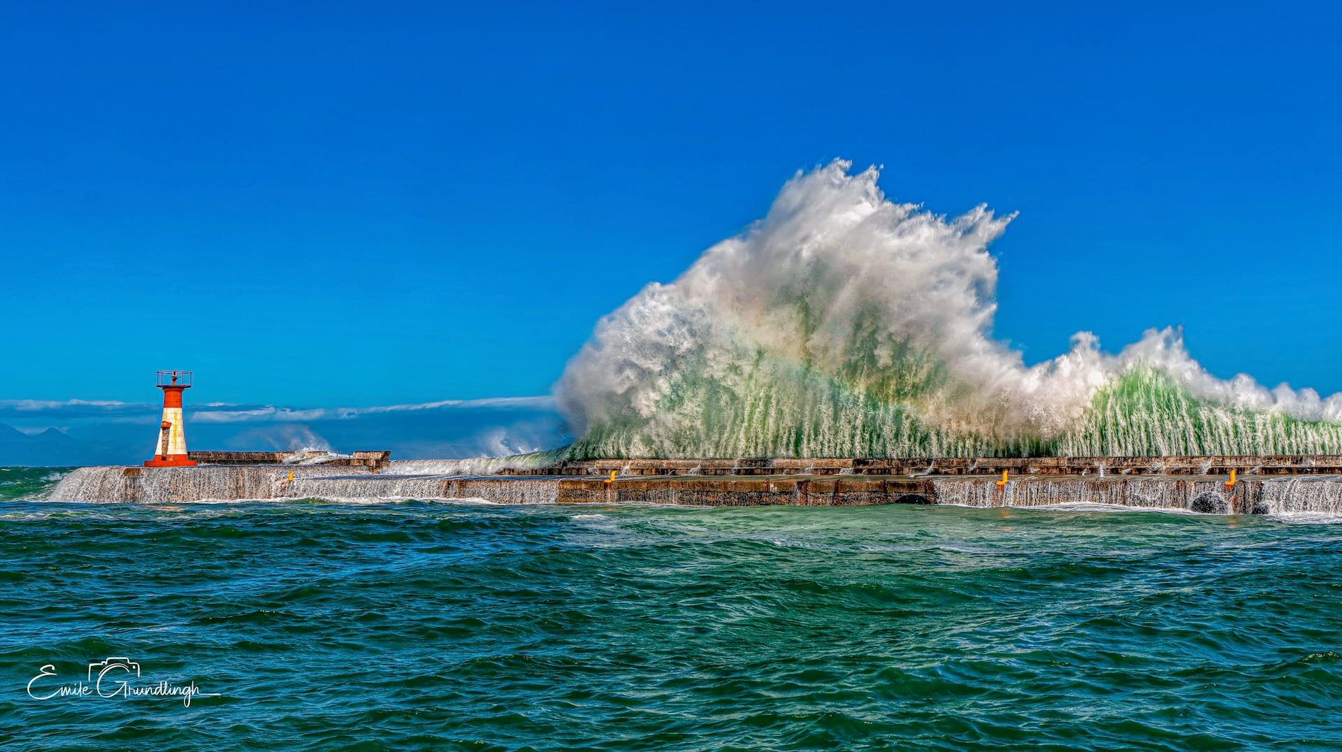Massive green wave cascading over Kalk Bay harbour wall under brilliant blue sky with red lighthouse, False Bay