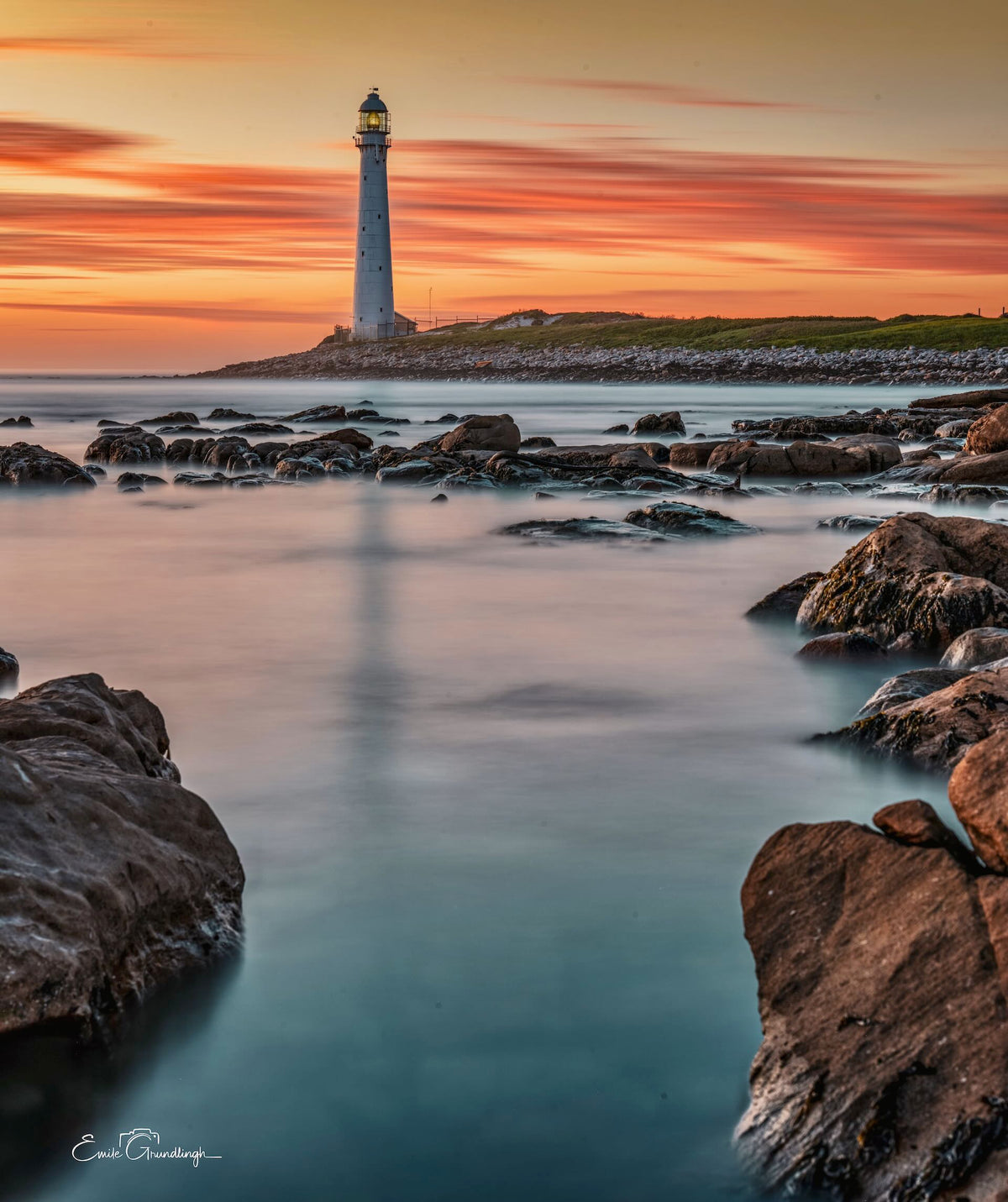 Slangkop Golden Hour – Slangkop Lighthouse framed by boulders at sunset with silky water and ghost reflection