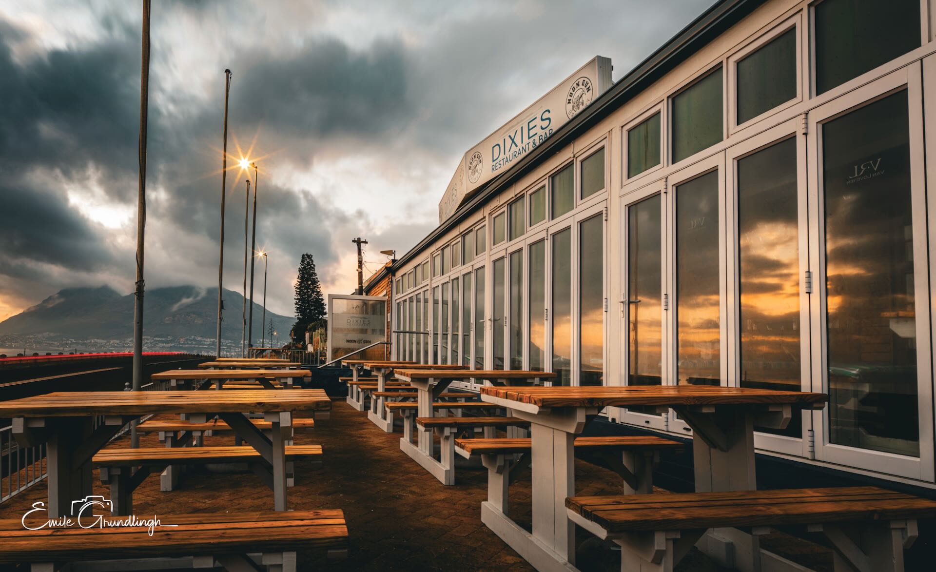 Empty terrace of Dixies Restaurant and Bar at Kalk Bay harbour at sunset with Table Mountain silhouette and storm clouds