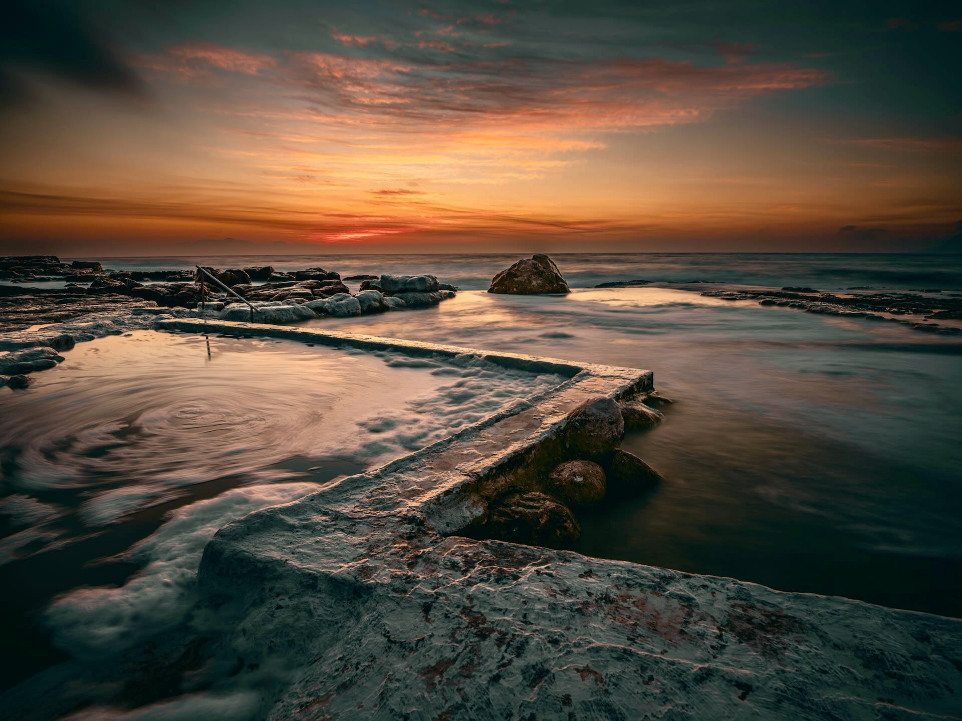 Kalk Bay tidal pool at sunset with silky swirling water over concrete walls and fiery orange sky