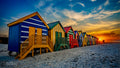 Close-up low angle view along Muizenberg beach huts toward a blazing golden sunset with vivid colours and dramatic wispy clouds