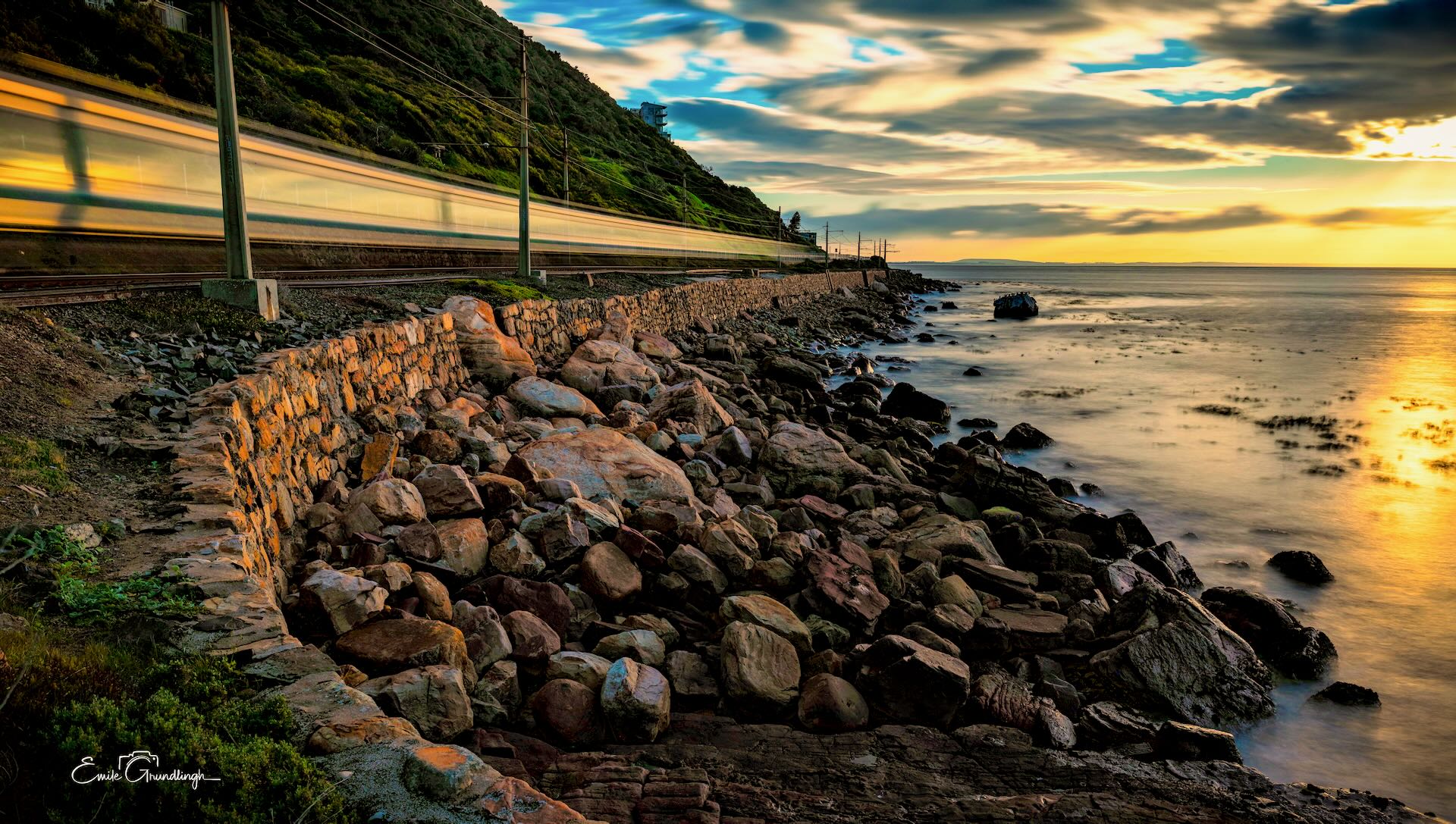 Motion-blurred train along the False Bay coastline at golden hour with rocky sea wall and silky ocean