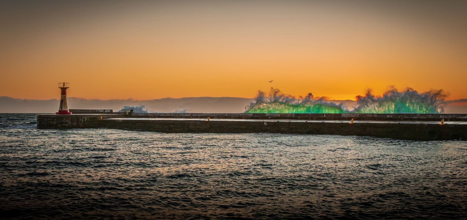 Massive emerald green wave exploding over Kalk Bay harbour wall at sunset with lighthouse in background