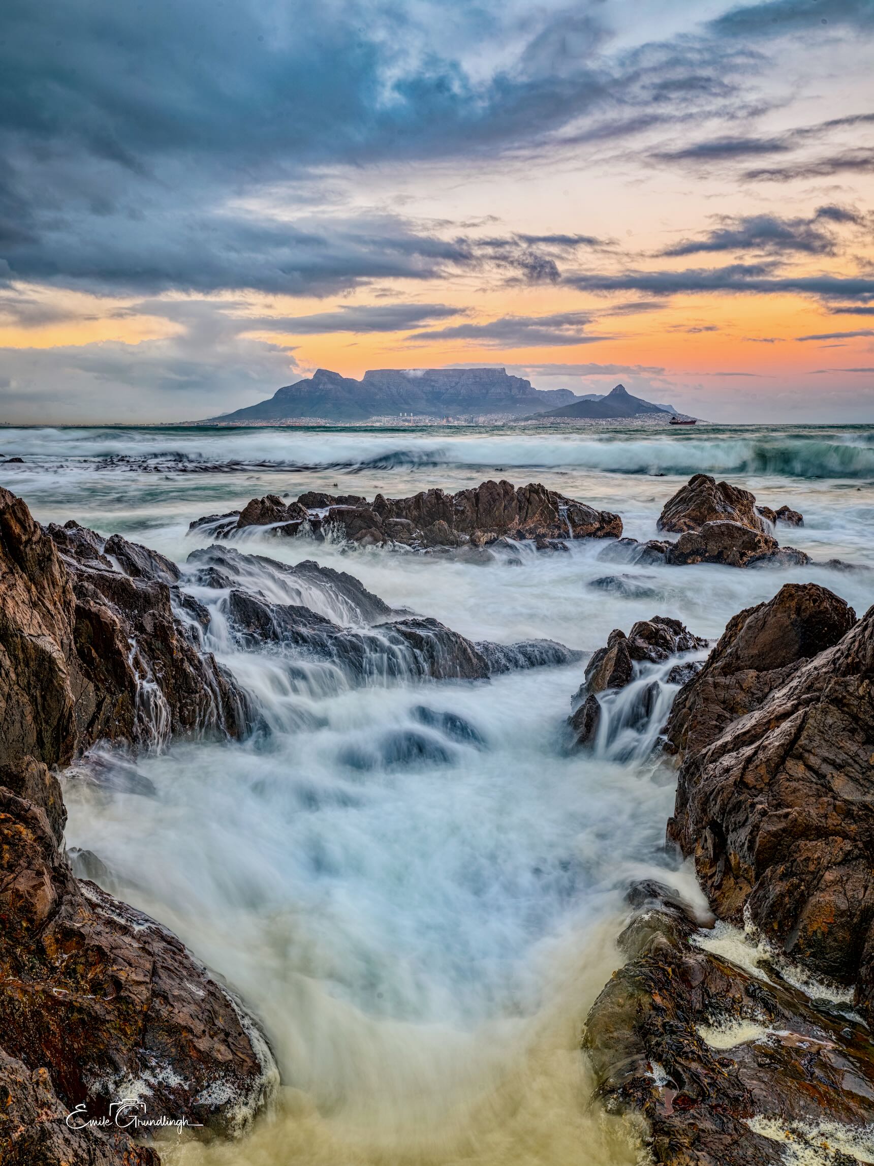 Table Mountain silhouette at sunset from Bloubergstrand with silky long exposure waves rushing through coastal rocks