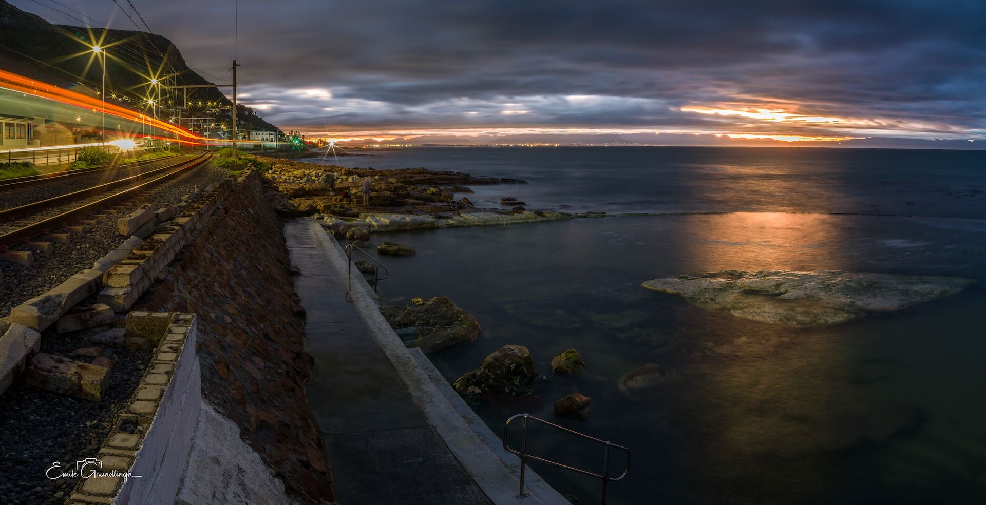 Dalebrook tidal pool at dusk with red and orange train light trails on coastal railway and False Bay sunset