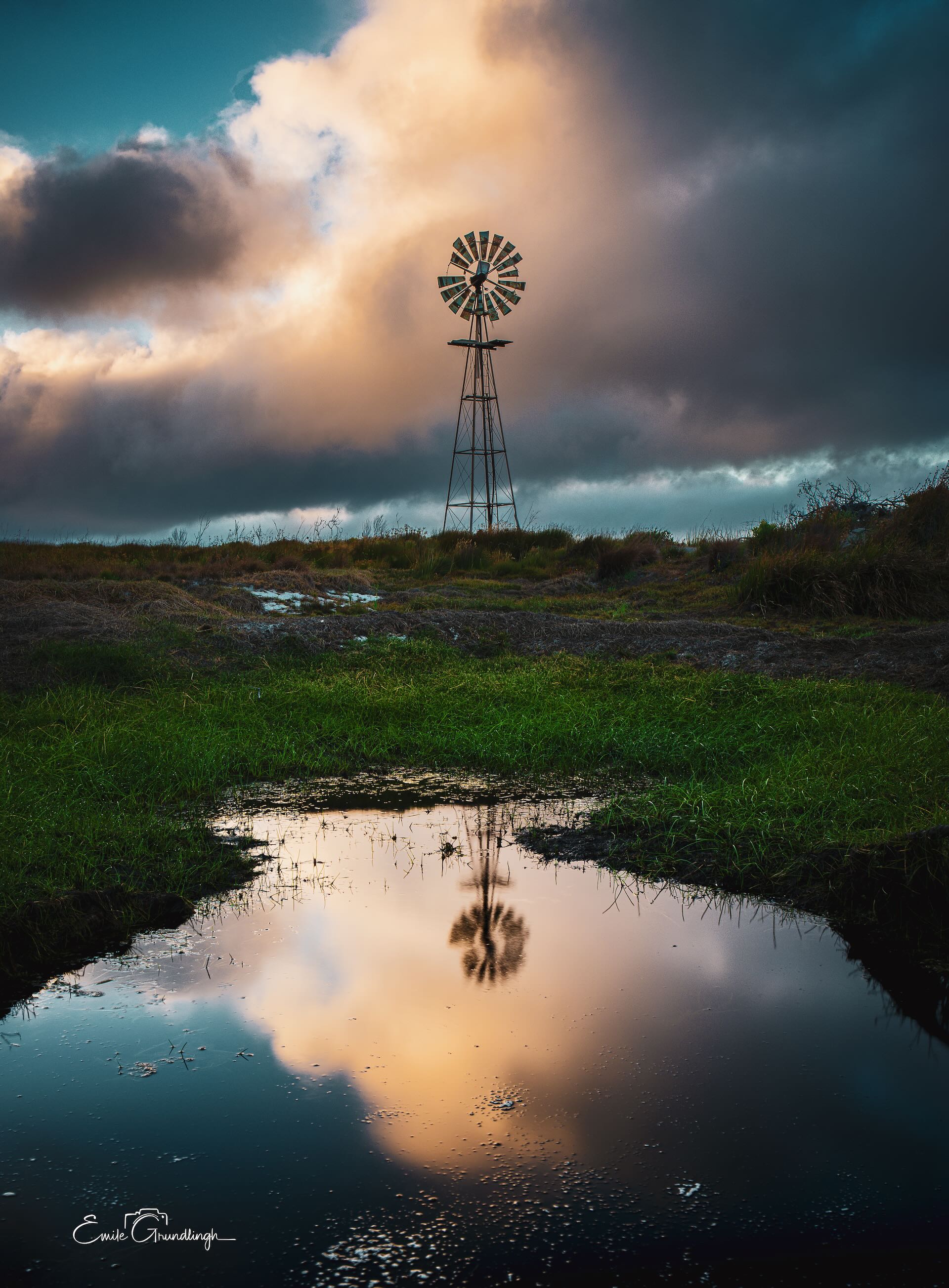 Lone windmill reflected in a still puddle against dramatic storm clouds and golden light at Cape Point