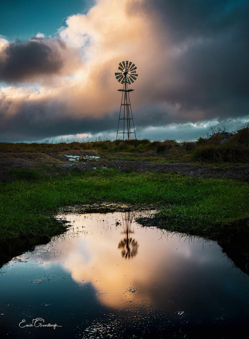 Windmill silhouetted against dramatic storm clouds reflected in a rain puddle on green farmland