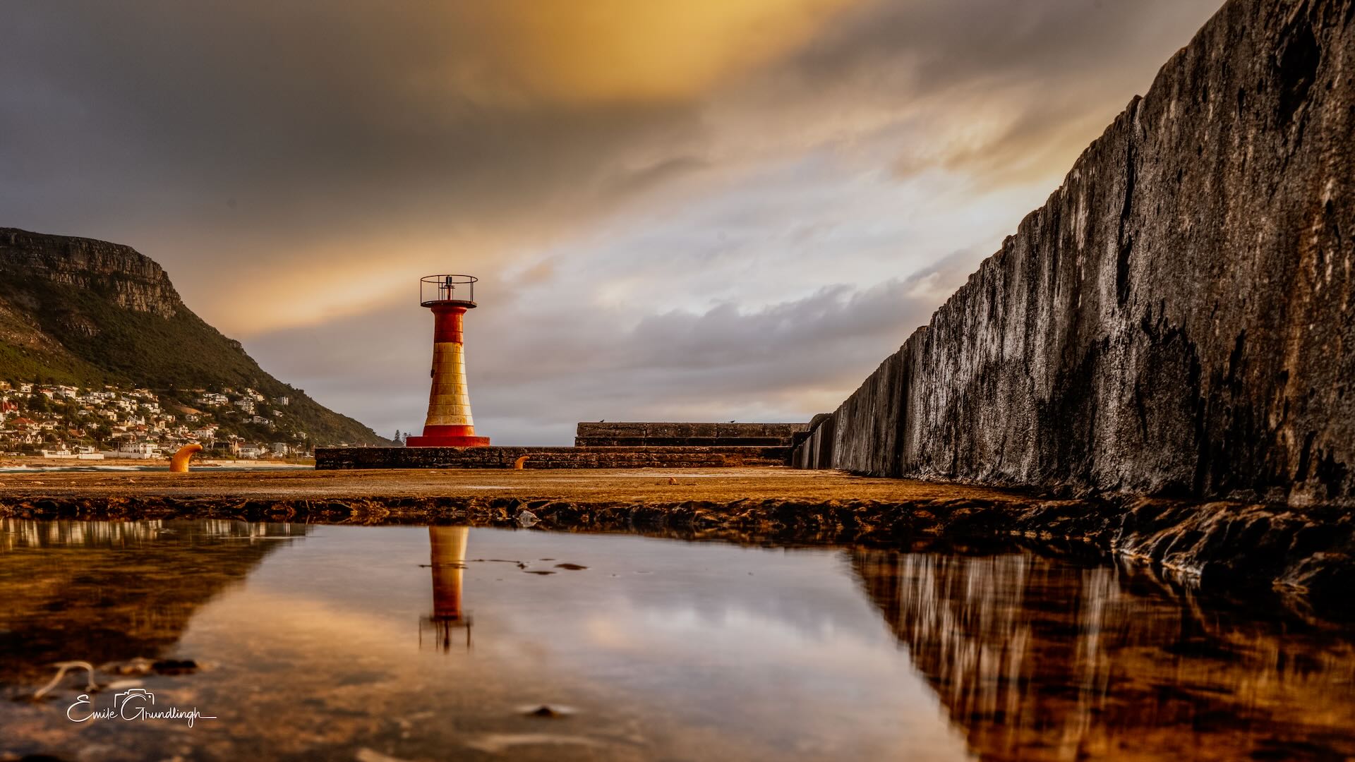 Kalk Bay lighthouse perfectly reflected in still rock pool on harbour wall with dark wall and mountain village backdrop
