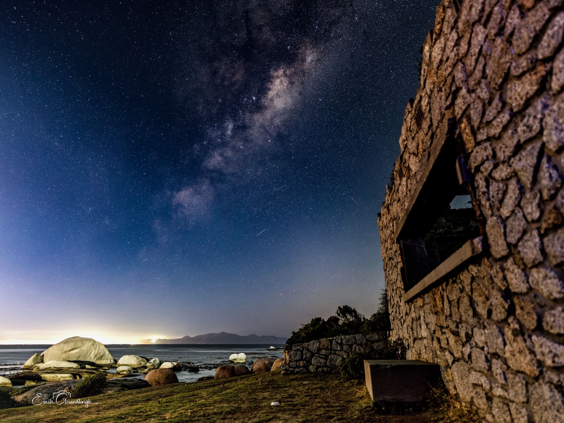 Milky Way arching over ancient stone ruins at Millers Point with granite boulders on False Bay shoreline at night