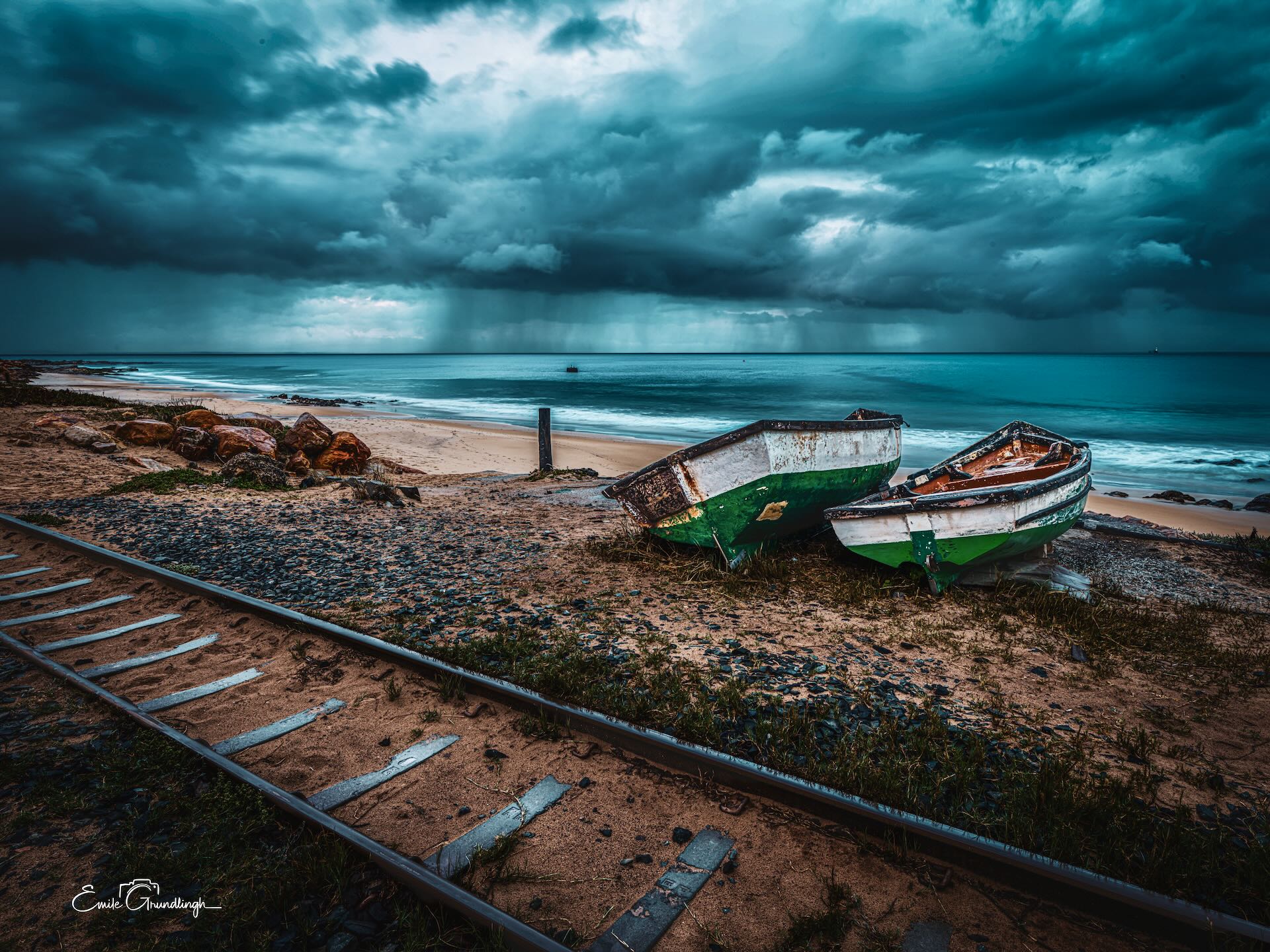 Two abandoned green fishing boats on boat launch rails at Makriels Beach with rain sweeping across False Bay under storm sky