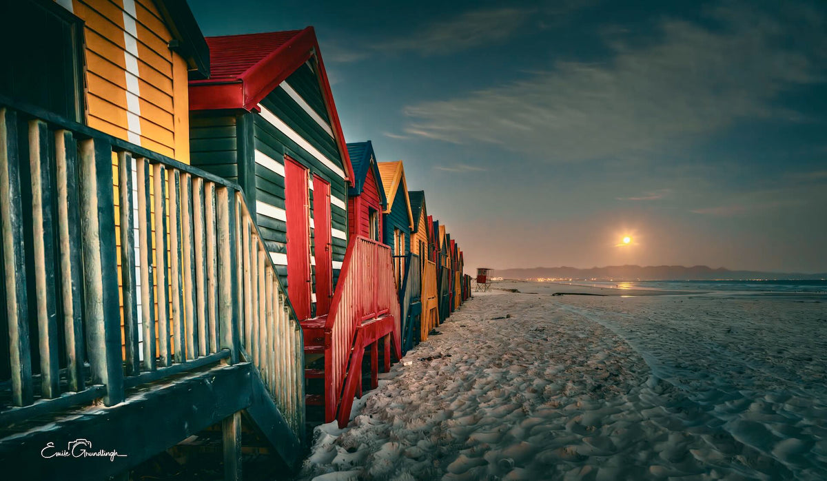Muizenberg beach huts glowing warmly at night with a full moon rising over False Bay under a deep blue sky