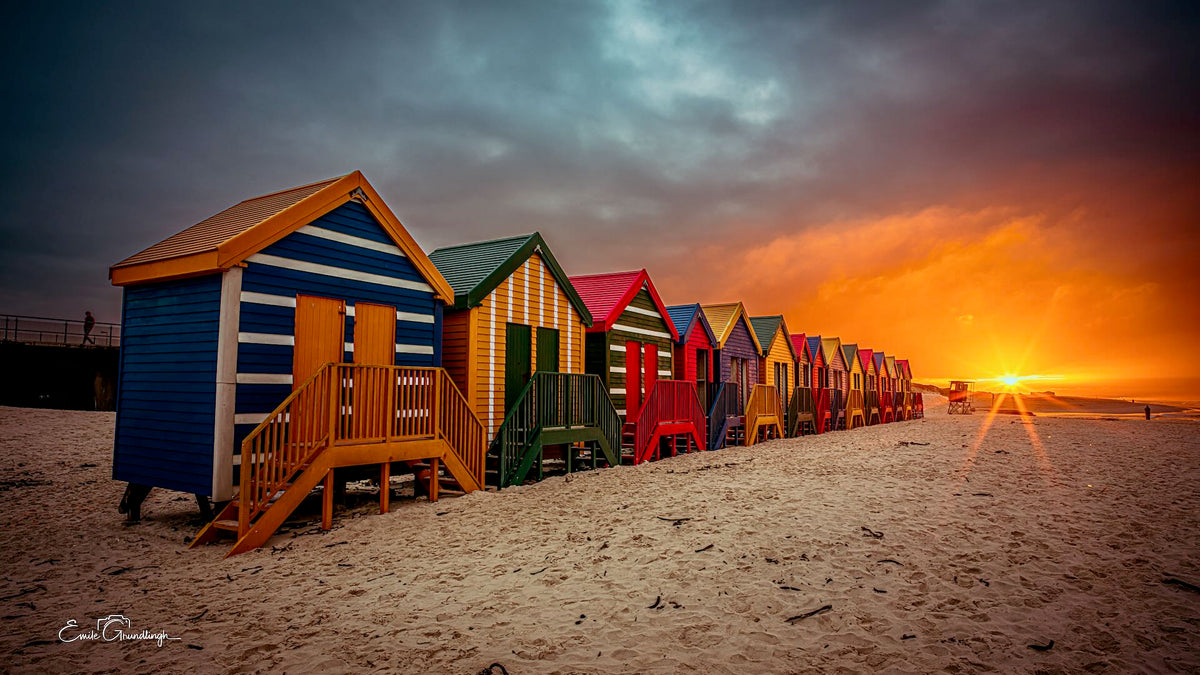 Row of colourful Muizenberg beach huts at sunset with a blazing orange sky and dramatic storm clouds overhead