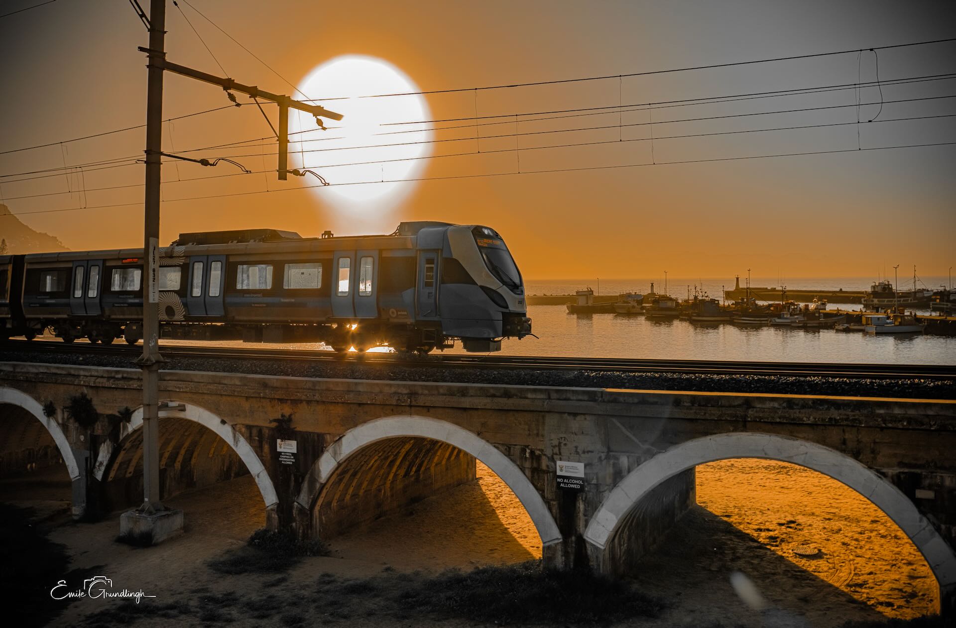 New Metrorail train crossing historic arched stone bridge at Kalk Bay harbour with glowing sun above against amber sunset sky