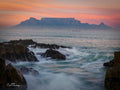 Table Mountain silhouetted against pink and orange sunset sky from Blouberg rocky shore with crashing waves