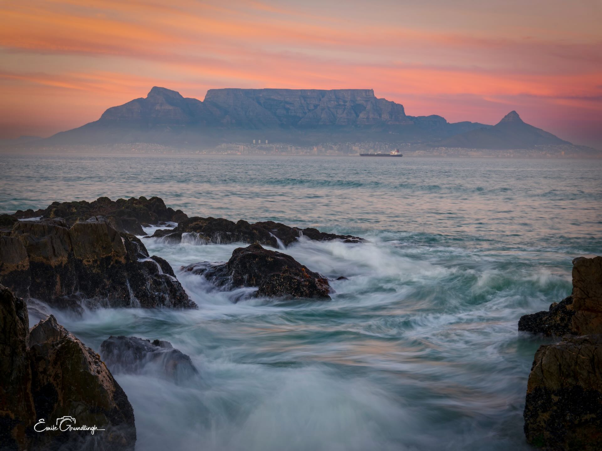 Table Mountain silhouetted against pink and orange sunset sky from Blouberg rocky shore with crashing waves