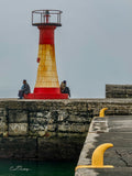 Two locals sitting on either side of Kalk Bay lighthouse on a grey misty day with yellow mooring bollards in foreground