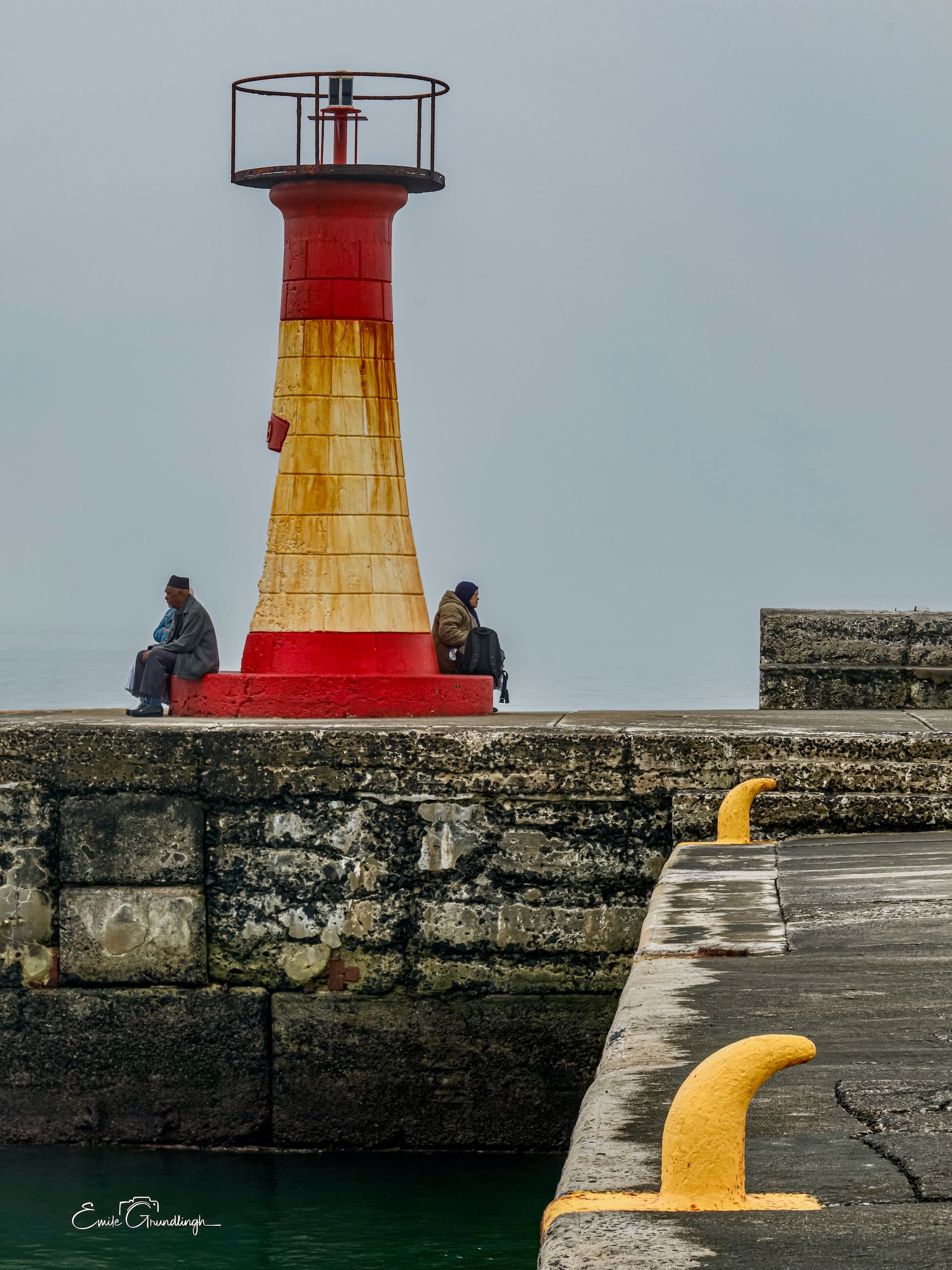 Two locals sitting on either side of Kalk Bay lighthouse on a grey misty day with yellow mooring bollards in foreground