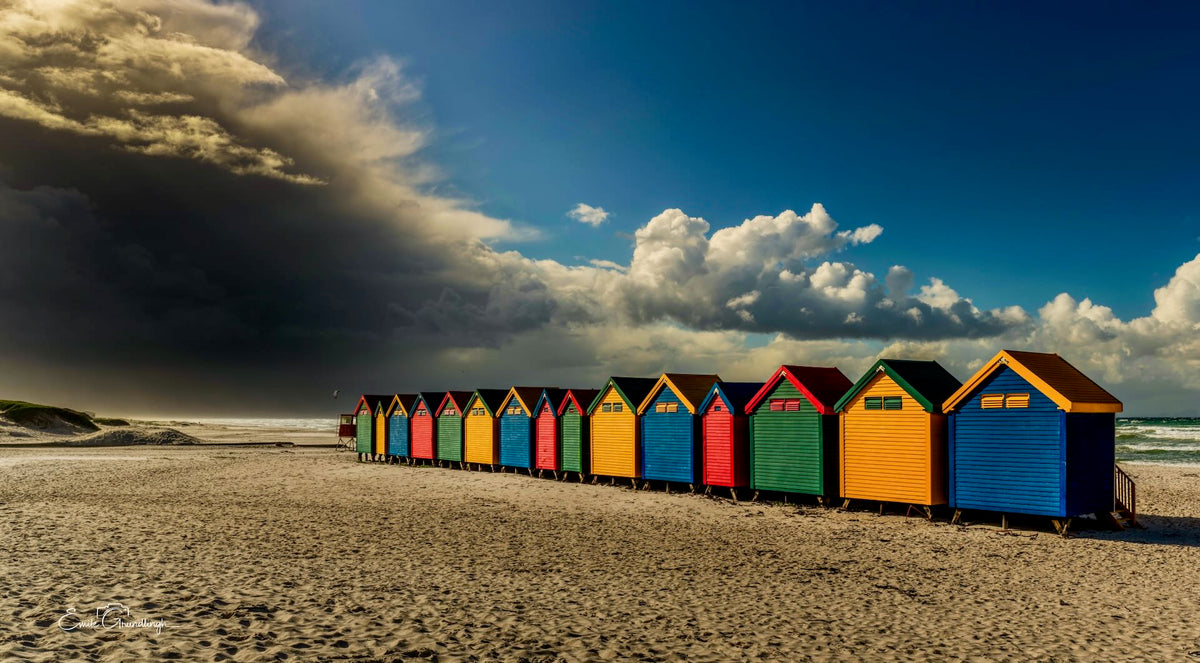 Row of colourful Muizenberg beach huts on empty sand with dramatic storm clouds rolling in from the left