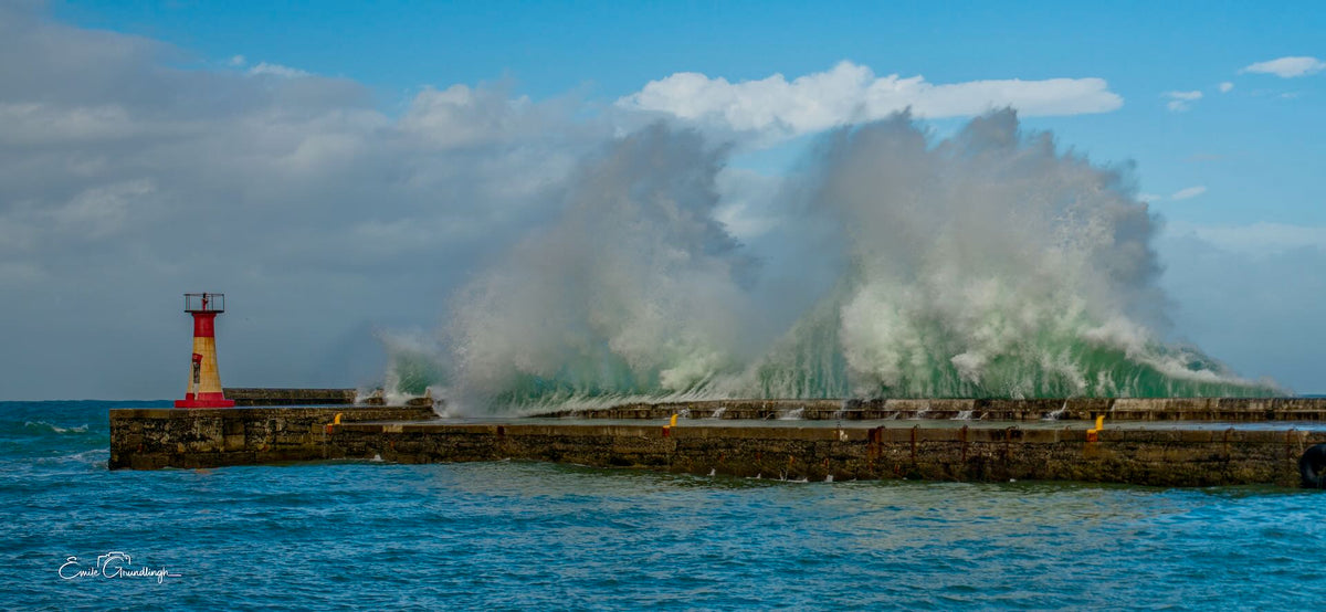 Giant wave crashing over Kalk Bay harbour wall with red and yellow lighthouse