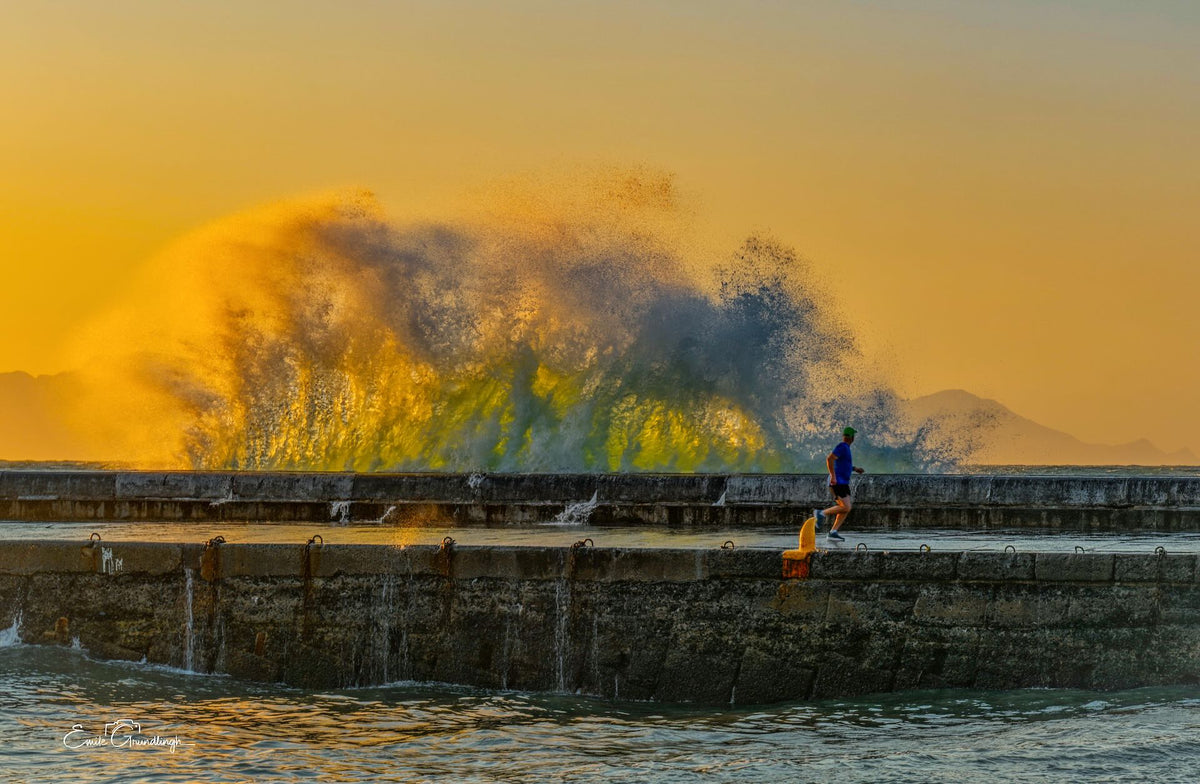 The Morning Runner – lone runner on Kalk Bay harbour wall with golden sunrise wave behind