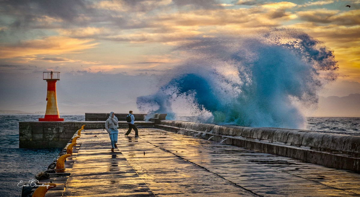 Two figures walking the Kalk Bay harbour wall as a massive turquoise wave curls dramatically behind them at sunset