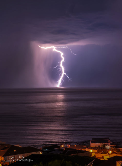 Single lightning bolt striking the ocean at night over a sleeping coastal town