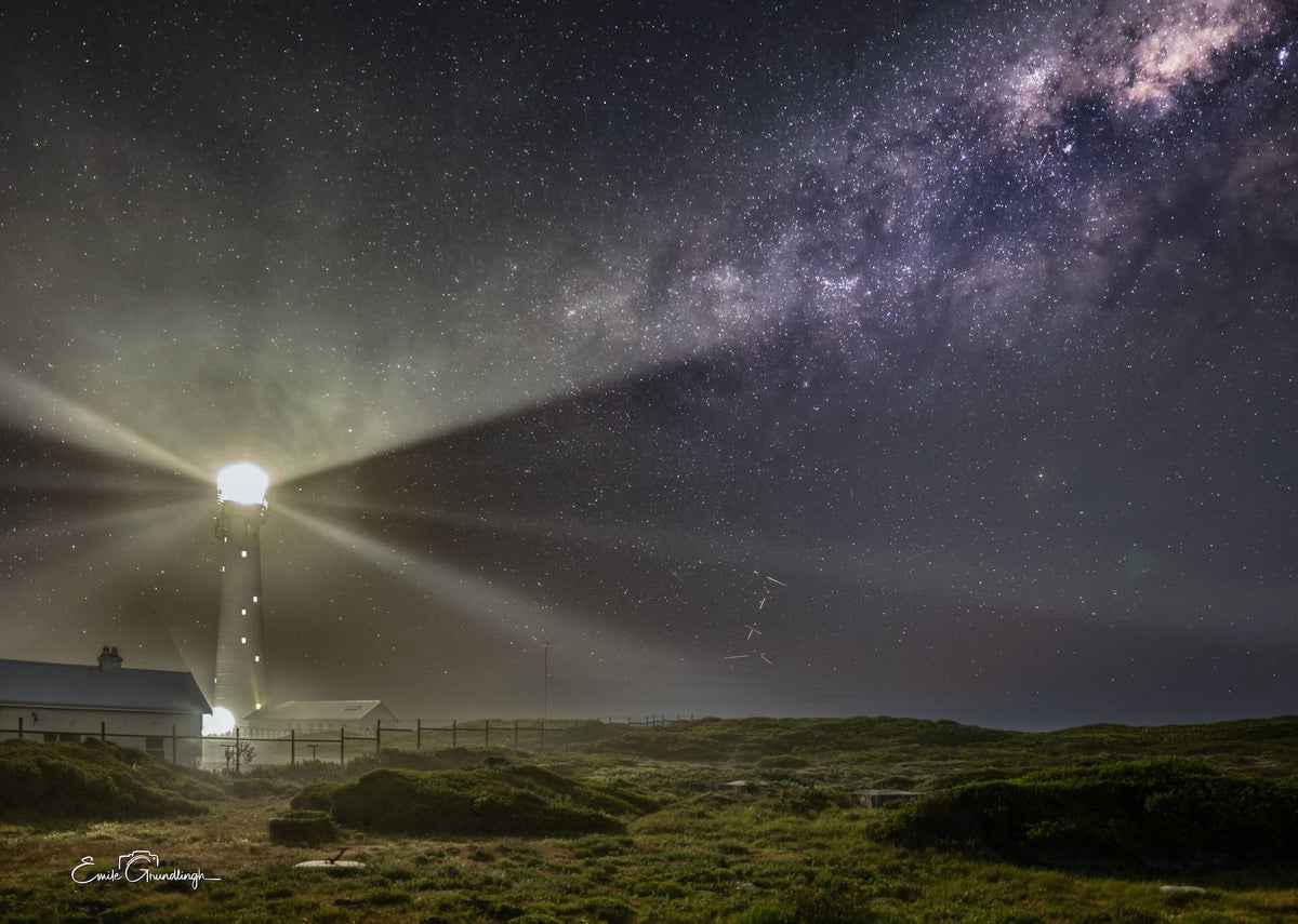 Slangkop Lighthouse casting beams beneath a diagonal Milky Way over the coastal landscape, photographed by Emile Grundlingh.