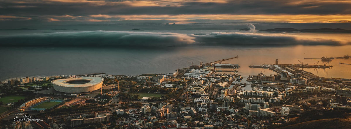 Panoramic sunset photograph of Cape Town showing the stadium and harbour, golden sky above and cloud rolling over the ocean, photographed by Emile Grundlingh.