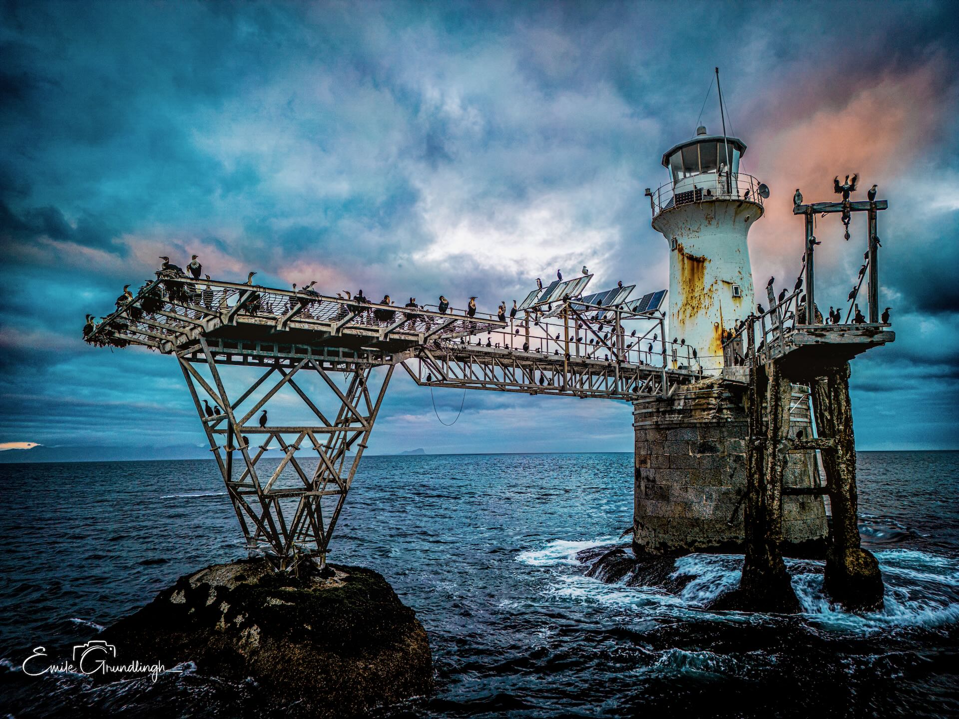 Roman Rock Lighthouse with steel gantry bridge covered in Cape cormorants under dramatic teal storm sky, False Bay