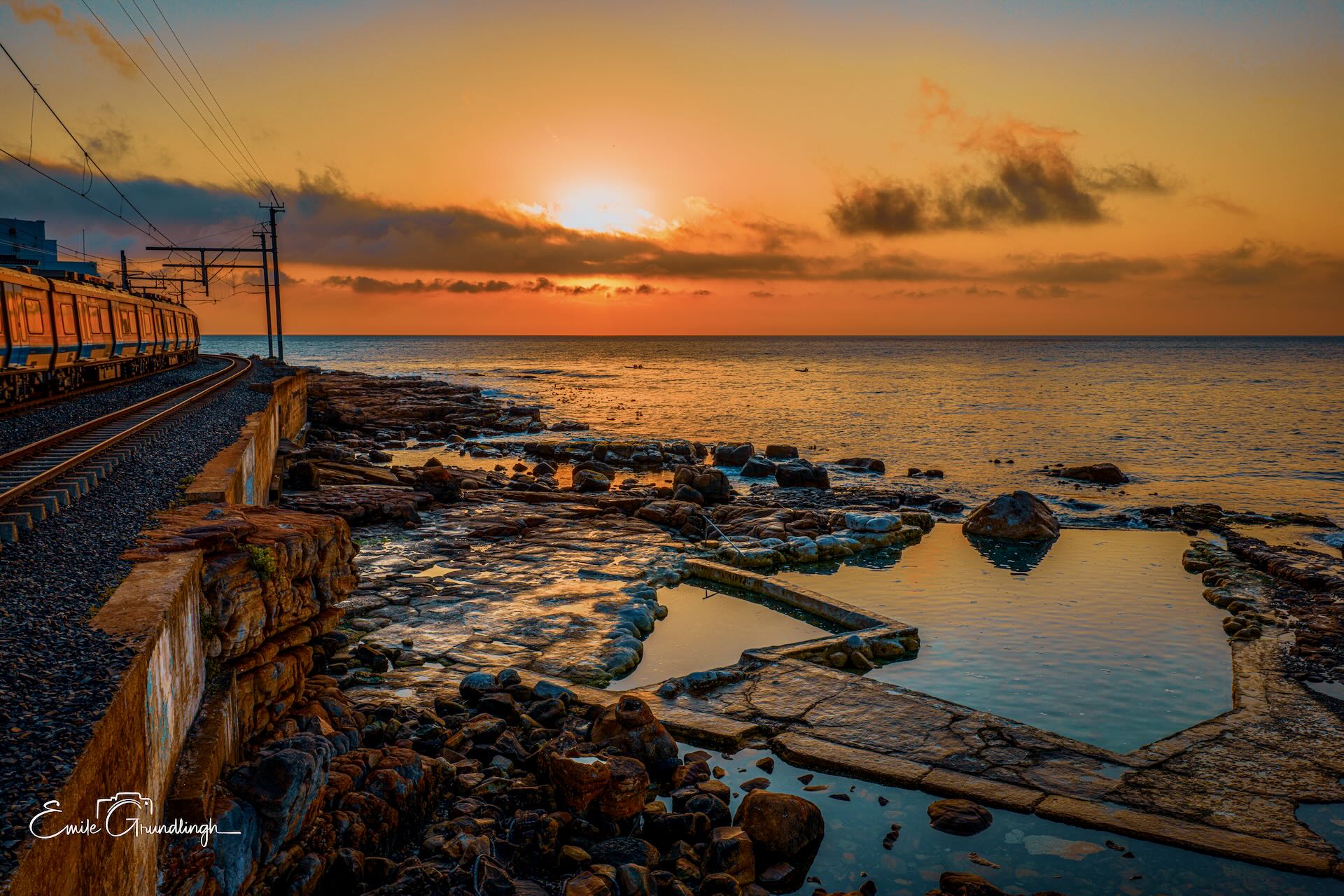 Wooleys Pool at blazing golden sunset with Metrorail coastal train passing alongside the tidal pool and False Bay beyond