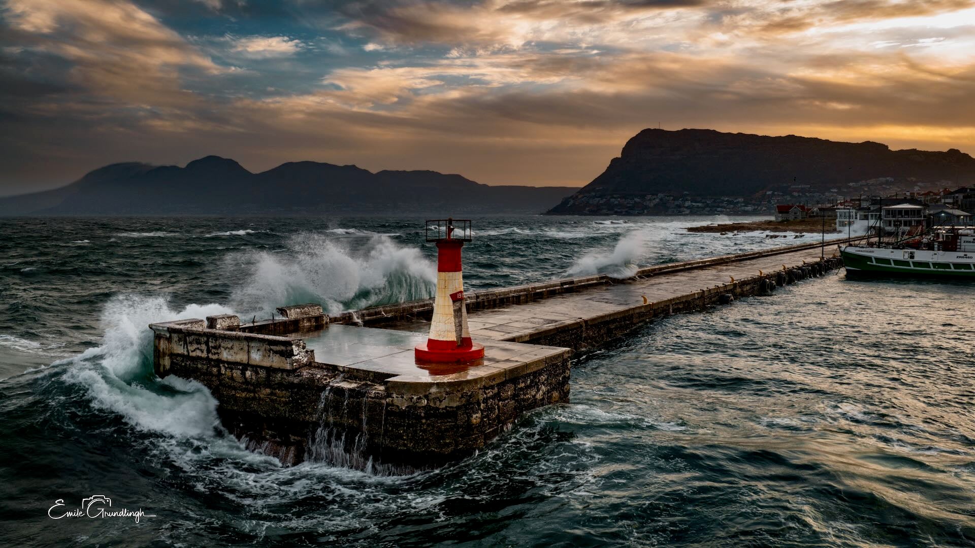 Aerial drone view of Kalk Bay harbour wall battered by storm swells at sunset with lighthouse and Hottentots Holland mountains